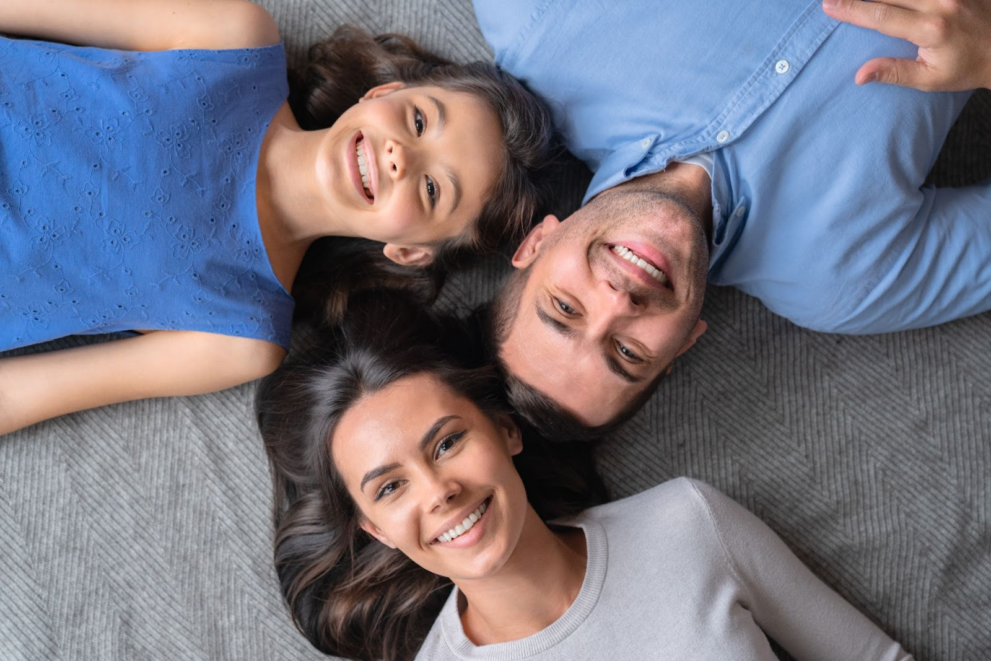 Happy family of three lying down with heads touching, illustrating a non arm's length transaction when buying a primary residence from a family member, involving equity gift, real estate agent, down payment, and self interest in investment property