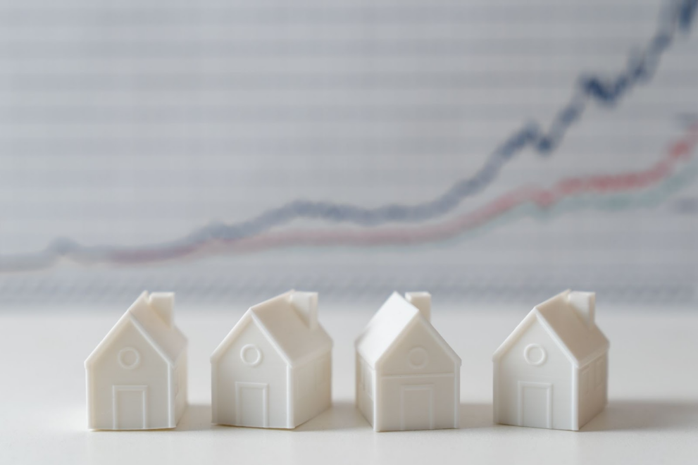 White toy houses representing a family house purchase with a blurred price chart in the background, illustrating non arm's length transaction, down payment considerations, and the importance of debt to income ratio and minimum credit score in the house hunting process.