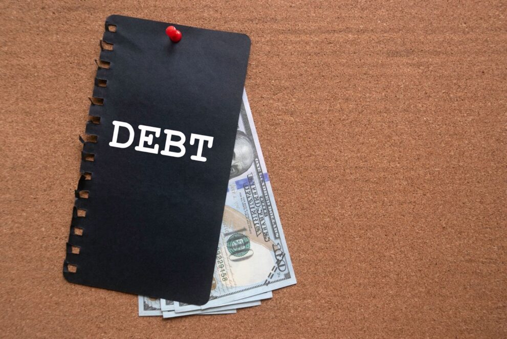 A black note with the word 'debt' is stuck on a notice board with some dollar currencies underneath it. This image represents the concept of debt-to-income ratio, where the amount of debt someone owes is compared to their income.