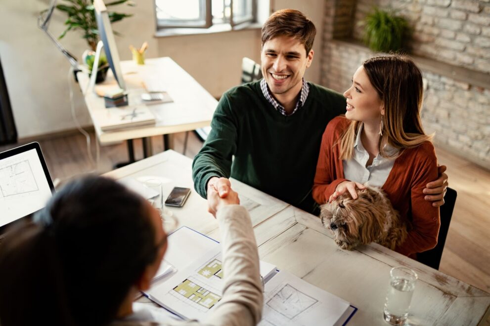 In an office, a man sits with his arm around a woman who is holding a dog while he reaches across a table to shake hands with another woman. On the table are real estate blueprints.