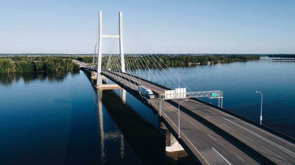 A truck drives across a large four-lane bridge that extends over a river. 